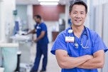 Male presenting nurse standing with their arms crossed in blue scrubs, smiling for the camera in a hallway of a hospital