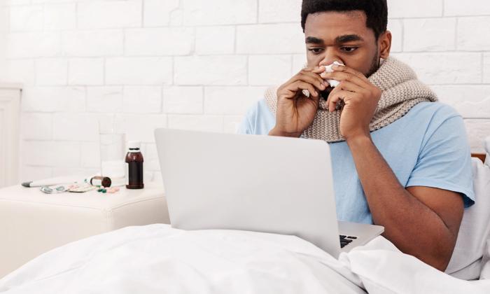 Man sitting in bed blowing his nose and looking at a laptop