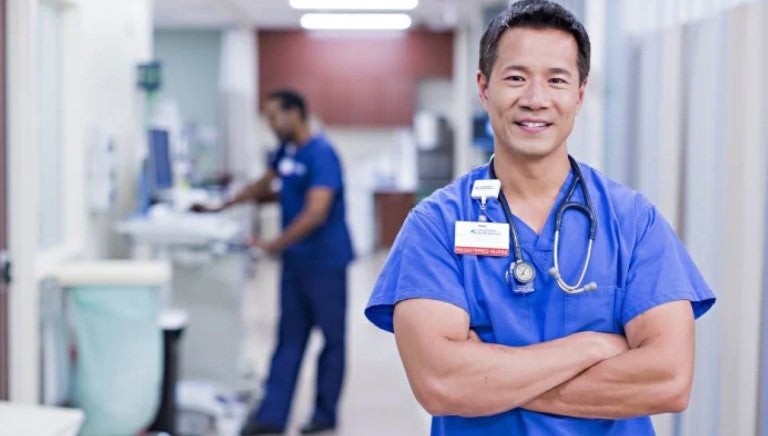 Male presenting nurse standing with their arms crossed in blue scrubs, smiling for the camera in a hallway of a hospital