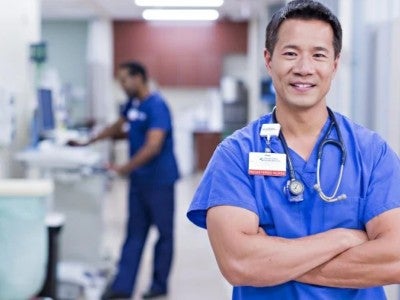 Male presenting nurse standing with their arms crossed in blue scrubs, smiling for the camera in a hallway of a hospital